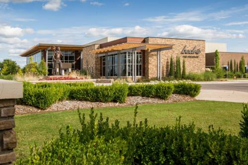 A modern building with stone and glass exterior, surrounded by greenery and a statue in front under a blue sky.