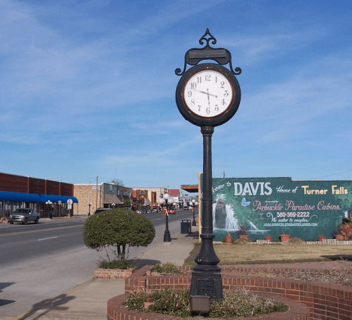 A vintage clock on a street corner, with a blue sky and buildings in the background, near a sign for Davis Cabins.
