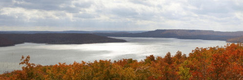 A serene lake surrounded by autumn foliage under a cloudy sky, reflecting soft light on the water's surface.