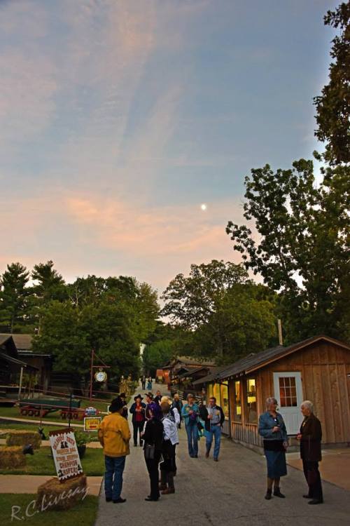A scenic view of a village at dusk, with people walking and chatting among wooden buildings and trees.