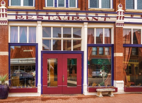 Front view of a restaurant with large windows, a maroon door, and decorative brickwork in a historic building.