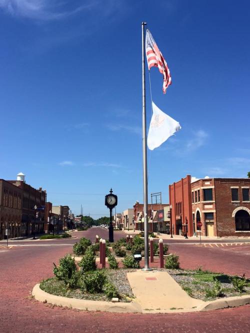 A small town square with a flagpole, American flag, and white flag, surrounded by brick buildings and a clear blue sky.