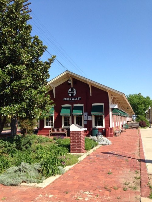 Historic red train station with green awnings, surrounded by greenery and a clear blue sky.