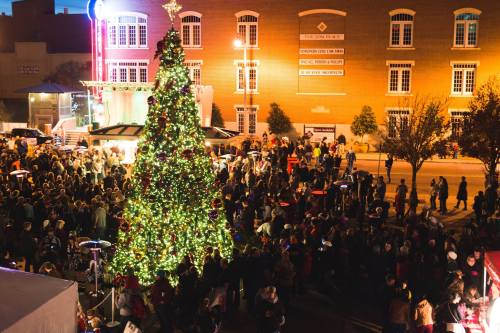 A large crowd gathers around a beautifully lit Christmas tree in a festive outdoor setting.