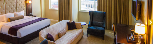 A spacious hotel room featuring a large bed, a cozy seating area, and a desk with a TV. Natural light from windows.