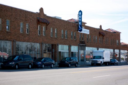 A brick hotel building with large windows and a blue "HOTEL" sign, parked cars and a white truck in front.