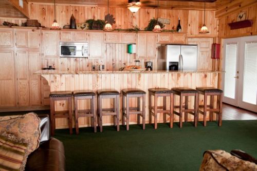A cozy wooden kitchen with a bar, stools, and modern appliances, featuring warm lighting and a green carpet.