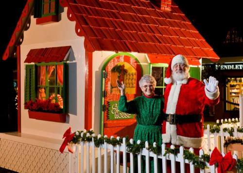 Santa Claus and a woman in a green dress stand outside a festive gingerbread house, waving cheerfully.