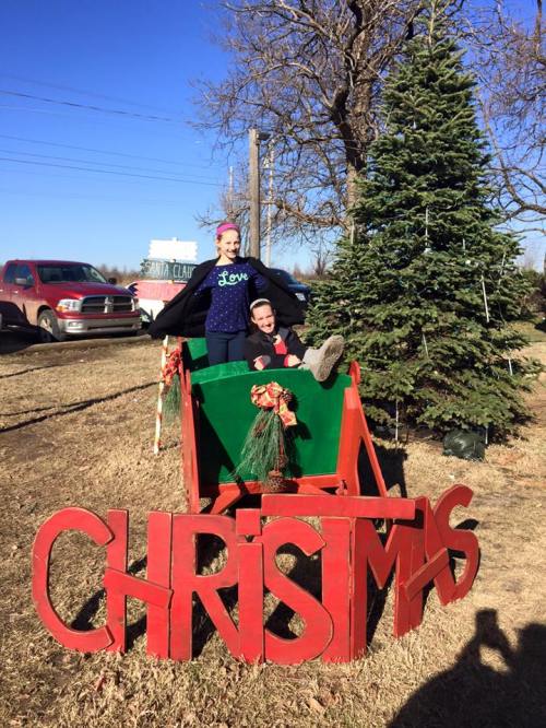 Two people playfully pose in a festive sleigh with a large "CHRISTMAS" sign, surrounded by a Christmas tree and a sunny backdrop.