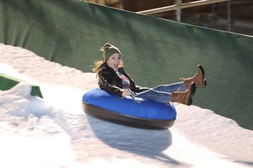 A person joyfully sliding down a snowy slope on a blue inner tube, wearing a beanie and winter clothing.