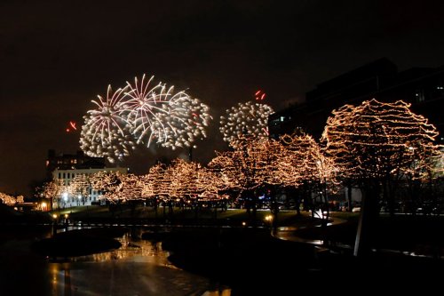 Fireworks illuminate the night sky above a park adorned with twinkling lights on trees, reflecting in the water below.