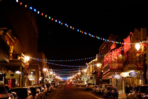 A festive street at night, adorned with colorful lights and decorations, lined with cars and shops.
