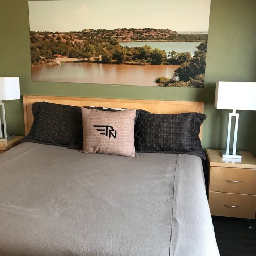A cozy hotel room with a bed, decorative pillow, and a scenic landscape painting of a lake on the wall.