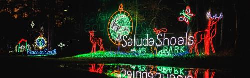 Colorful holiday lights display at Saluda Shoals Park, featuring a globe, deer, and festive messages reflected in water.