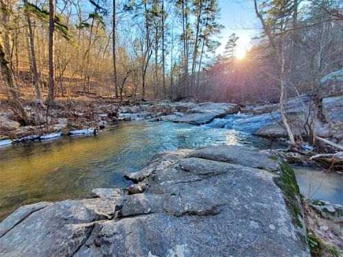 A serene river flows over rocks, surrounded by trees, with the sun setting in the background.