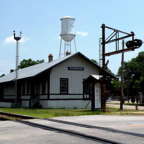 Historic train station in Pinewood with a water tower and railroad crossing signals under a clear blue sky.