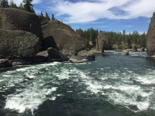 A river flows between large rocks, surrounded by lush green trees and a blue sky with scattered clouds.