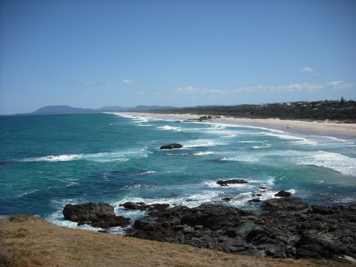A scenic view of a coastline with rocky shores, gentle waves, and a sandy beach under a clear blue sky.