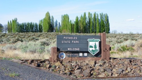 Welcome sign for Potholes State Park, surrounded by shrubs and trees under a clear blue sky.