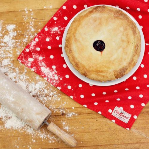A freshly baked pie on a white plate, resting on a red polka dot cloth, with a rolling pin and flour scattered nearby.