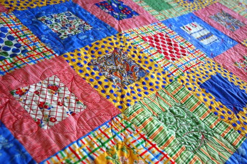 A colorful quilt featuring various patterns in squares, including polka dots, stripes, and floral designs.