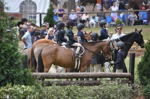 A group of young riders on horseback prepare for a competition, with spectators and horses in the background.
