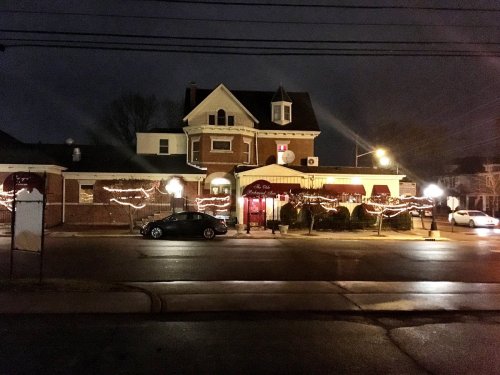 A charming restaurant illuminated with festive lights at night, featuring a Victorian-style building and parked cars.