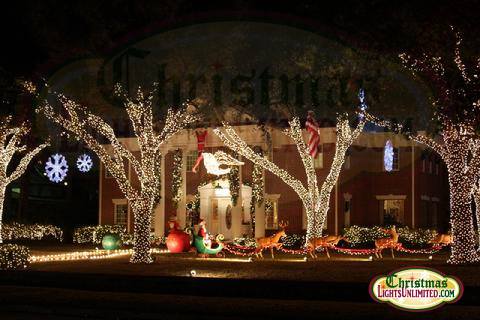 A festive house decorated with colorful Christmas lights, featuring a reindeer, sleigh, and snowflakes in the trees.