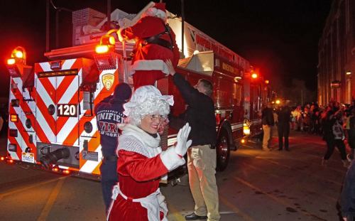 Santa and Mrs. Claus wave as they arrive on a fire truck, surrounded by a festive crowd at night.