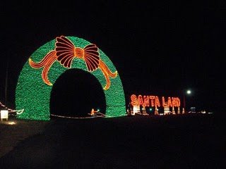 A brightly lit arch with a bow and the words "SANTA LAND" illuminated in festive colors at night.