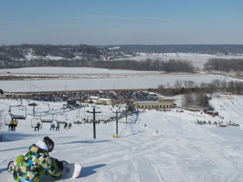 A snowy ski slope with a snowboarder in colorful gear, overlooking a parking lot and scenic winter landscape.