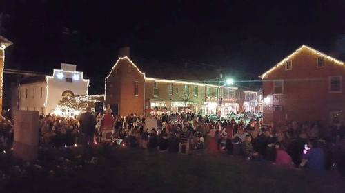 A large crowd gathers in a town square at night, illuminated by festive lights on nearby buildings.
