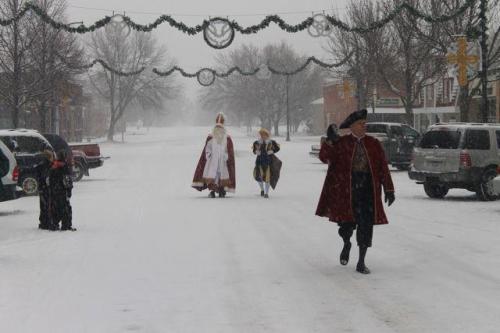 A snowy street scene with people in festive costumes walking, surrounded by holiday decorations and snow-covered trees.