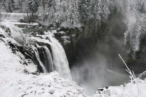A snowy waterfall cascades down rocky cliffs, surrounded by frosted trees and mist rising from the water.