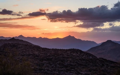 Sunset over rugged mountains, with vibrant colors in the sky and clouds scattered above the horizon.