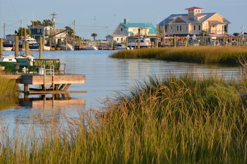 A serene waterfront scene with boats, docks, and grassy banks under a clear sky. Charming buildings line the shore.