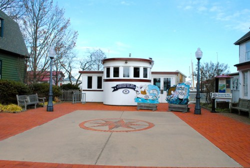 A waterfront building with a circular design, surrounded by benches and a compass rose on the ground. Clear blue sky.