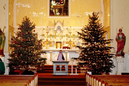 A decorated church interior with two Christmas trees, a central altar, and religious statues. Warm, festive atmosphere.