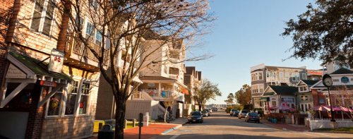 A charming street lined with shops and trees, leading to a waterfront in a sunny, coastal town.