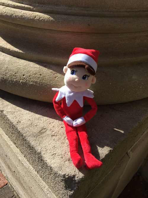 A plush elf doll in a red and white outfit sits on a stone surface, smiling in the sunlight.