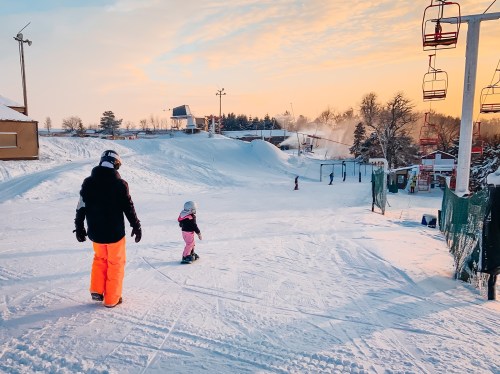 A person and a child walk on a snowy slope, with ski lifts and trees in the background under a colorful sky.
