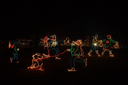 Colorful holiday lights depict people and a dog walking in a festive scene at night.