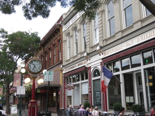 Historic street scene featuring a vintage clock, shops, and outdoor seating with trees in the background.