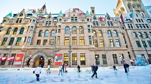 Historic building with ornate architecture, ice skating rink in front, and people skating on a sunny day.