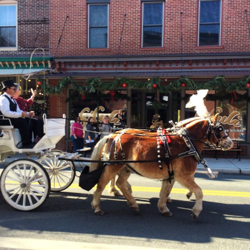 A horse-drawn carriage with passengers rides down a festive street decorated for the holidays.