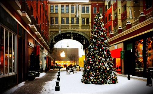 A festive alleyway with a large decorated Christmas tree, shops, and snow, leading to an Apple store in the background.