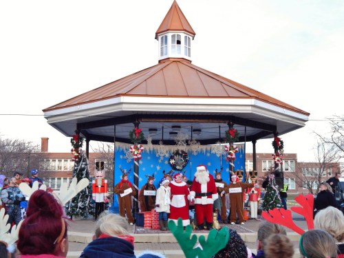 A festive stage with Santa and performers in holiday costumes, surrounded by decorations and an audience.