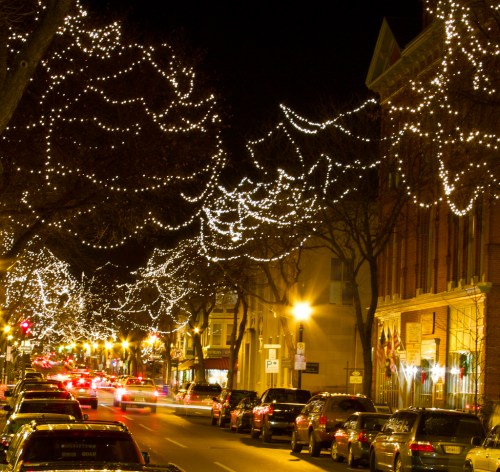 A festive street at night, lined with cars and illuminated by twinkling holiday lights in the trees.