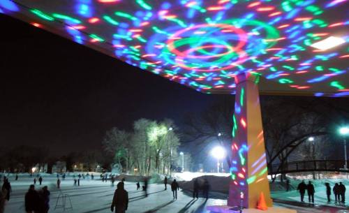 Colorful lights reflect on the ice as people skate at night, surrounded by trees and illuminated by bright lamps.