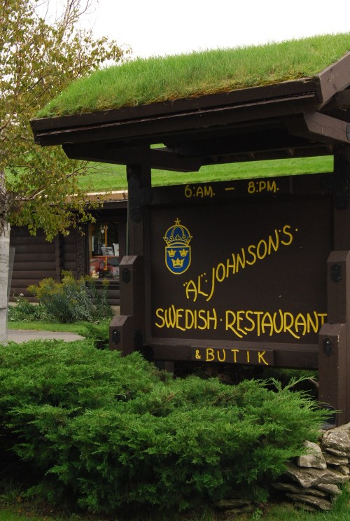 Sign for Al Johnson's Swedish Restaurant & Butik, featuring a green roof and surrounded by greenery. Open 6 AM - 8 PM.